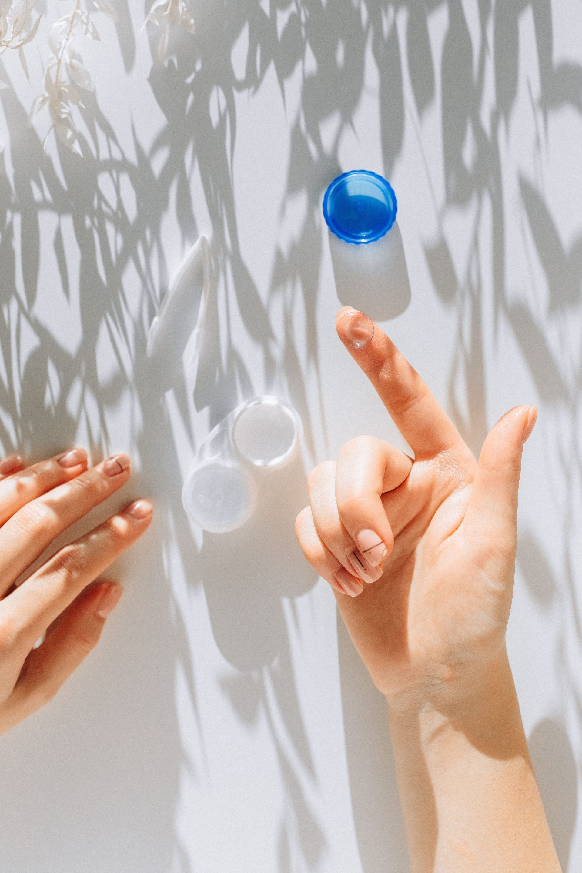 Gros plan sur des mains tenant une lentille de contact près d'un étui, éclairées par une douce lumière du soleil sur une surface blanche.